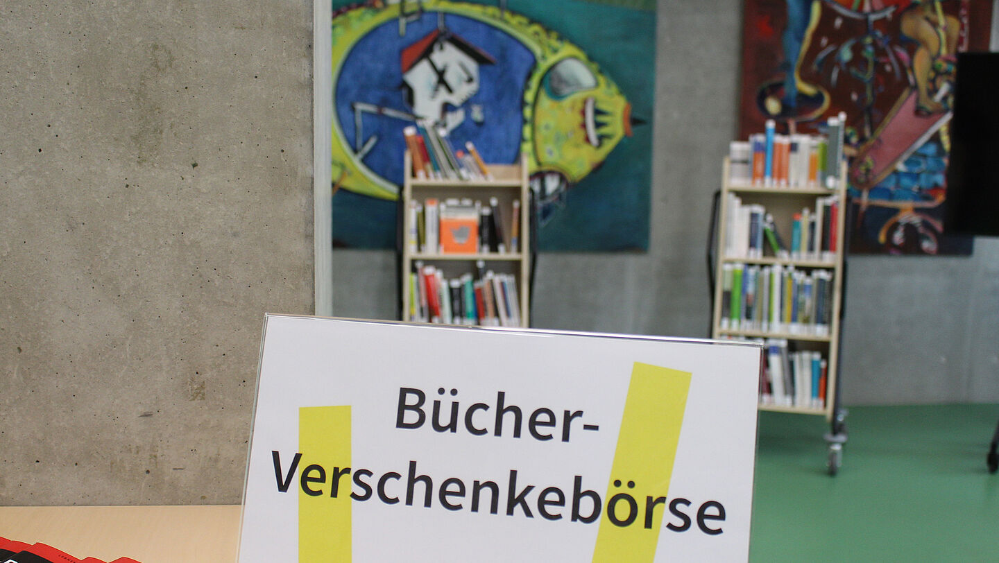 Schild "Bücher-Verschenkebörse" und dahinter Bücherwagen im Bibliotheksfoyer