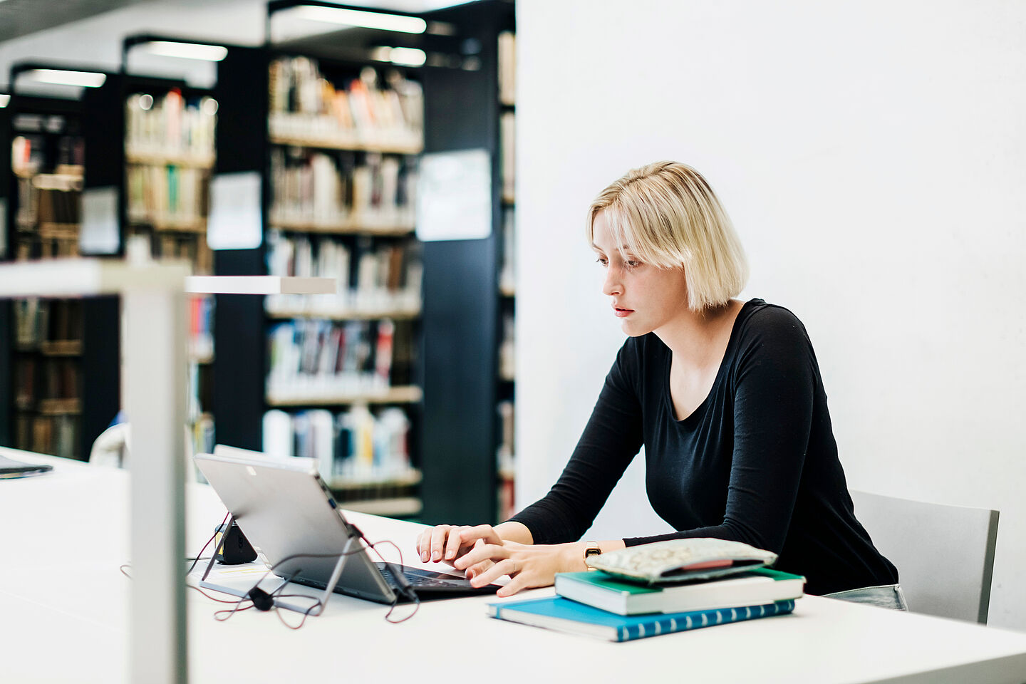 Eine blonde Frau sitzt an einem Tisch vor einem Laptop. Links von ihr liegen Bücher auf dem Tisch. Im Hintergrund sind Bücherregale zu sehen.