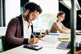 Student mit Stift, Taschenrechner und Laptop sitzt in der Bibliothek - im Hintergrund eine Studentin
