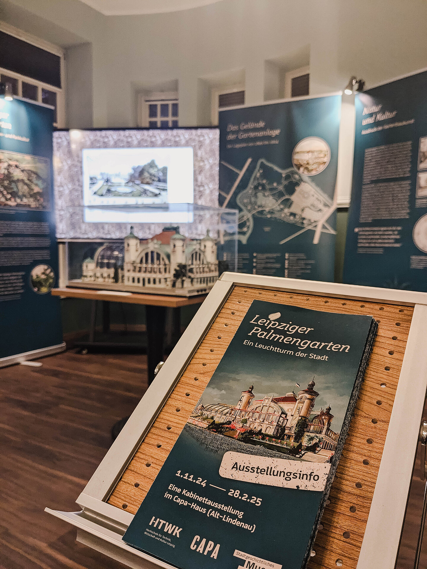 Blick in die Ausstellung mit dem Modell des Gesellschaftshauses (hinten). Blick in einen Raum über eine Broschüre hinweg