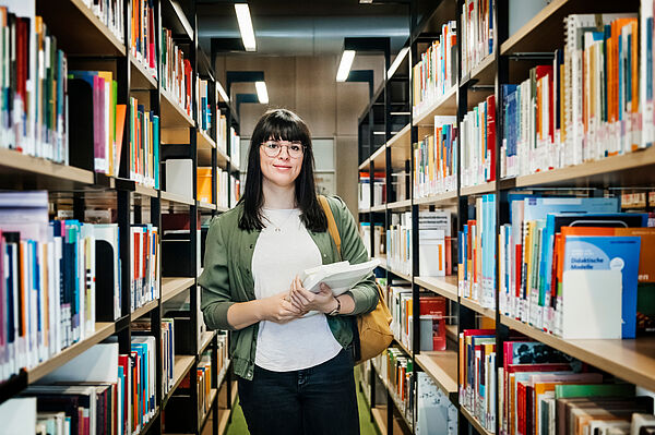 Eine Frau mit Unterlagen in der Hand steht zwischen Bücherregalen in der Bibliothek.