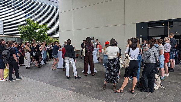 Ausstellungseröffnung Museologie-Reihe am 24. Juni 2025 Gruppe von Menschen steht auf einem Platz zwischen Gebäuden, manche an Stehtischen