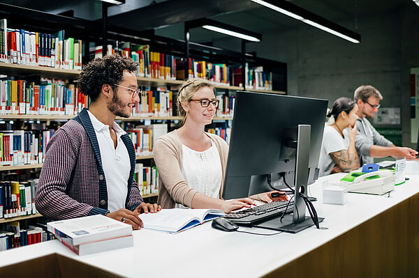 Zwei Studierende stehen vor einem Recherche-Rechner in der Bibliothek
