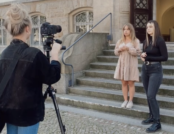 zwei Frauen im Hintergrund, im Vordergrund links Rückansicht einer Frau, die die beiden anderen mit einer Kamera auf Stativ filmt 